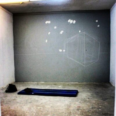 Installation art by contemporary artist Isaac Cohen. the image features an empty room with a faint cube and horizon drawn on the wall. on the floor there is a mattress and a bag